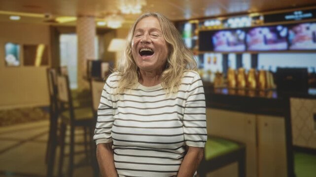 Senior woman with open mouth laughing and clapping hands at hotel bar counter with beer taps and tv screens; joy.