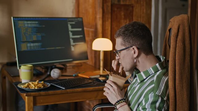Medium handheld shot of young Caucasian freelance programmer coding on desktop computer and eating noodles from box while sitting at table, working from home