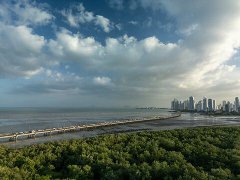 Panama city skyline rising under a cloudy sky during sunset, with the corredor sur highway extending across the ocean, showing busy traffic and lush mangroves in the foreground - stock photo