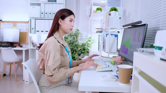 Professional businesswoman in office typing on computer keyboard at modern workplace, reviewing finance report with data analysis and chart on monitor, corporate desk workflow focused work