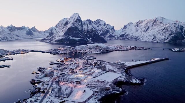 Aerial view of the snow-covered village of Reine in Lofoten with majestic mountains and calm fjords under a soft twilight sky in Nordland, Norway.