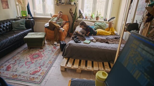 Handheld shot of Black freelance developer putting on headphones coding on computer while two Caucasian roommates distracting him in background in rented apartment