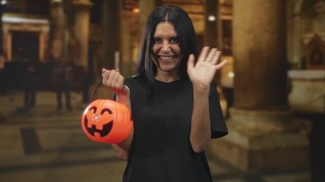 Woman smiling broadly and waving hand while holding orange pumpkin bucket in ornate building; playful joy.