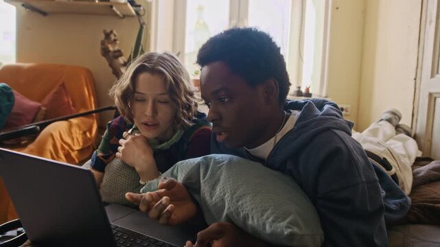 Handheld shot of two diverse cheerful gen Z freelancers discussing working tasks while lying on bed and using laptop in shared apartment