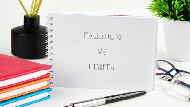 Words Freedom versus Limits clearly appear on a white spiral notebook. Placed on a clean desk with colorful books, a pen, eyeglasses, a plant, and a diffuser