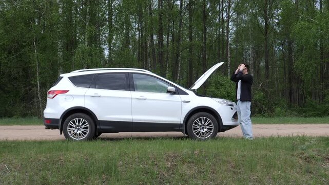 Stressed man standing beside a broken white suv with its hood open on the side of a rural road, frustrated by the unexpected vehicle engine failure and seeking assistance