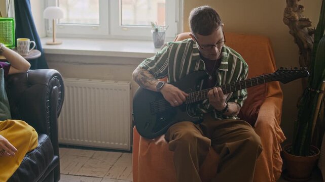 Long handheld shot of young Caucasian woman enjoying male roommate playing guitar and distracting Black gen Z freelancer working on laptop on bed, living in shared apartment
