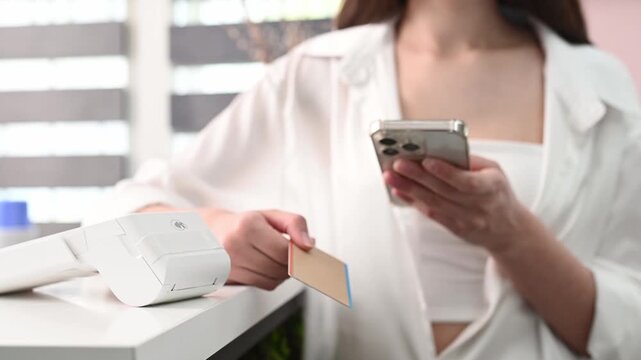 wireless payment terminal in beauty salon, enter PIN code,  closeup of hands unrecognizable. Girl pays with card through POS terminal and scrolls through feed on smartphone