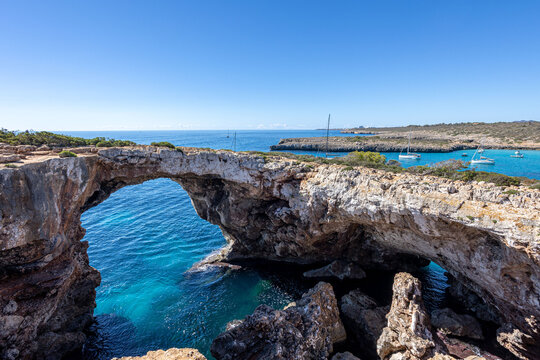 Scenic rocky arch forming a natural bridge above clear blue water at Cala Varques beach on Mallorca east coast under bright sunny sky