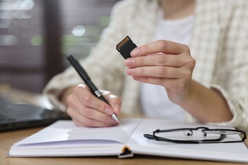 Fototapeta premium Woman with SD memory card taking notes at wooden table indoors, closeup