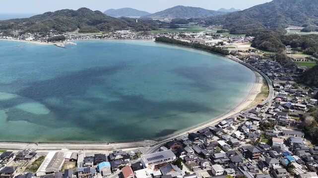 Aerial view of Itoshima Peninsula featuring the curved sandy beach, coastal houses, and lush green hills under a clear sky in Itoshima, Fukuoka, Japan.