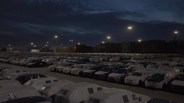 Night view of large car storage lot at port under stormy sky and industrial lights