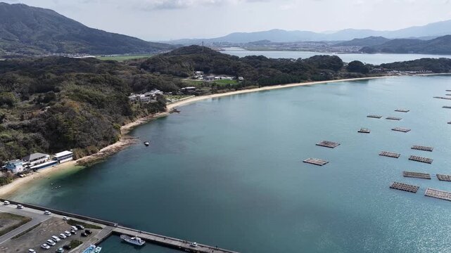 Aerial view of Itoshima Peninsula featuring floating oyster farms, a concrete pier with boats, and forested hills along the coastline in Itoshima, Fukuoka, Japan.