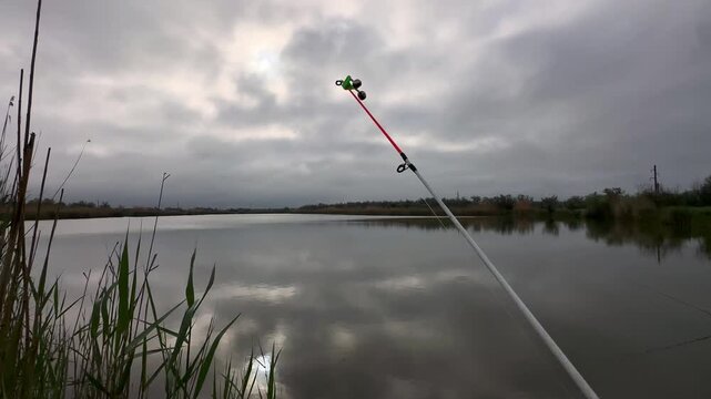 Fishing rod shaking during fish bite near lake under cloudy sky. Concept of recreational fishing and outdoor activity.