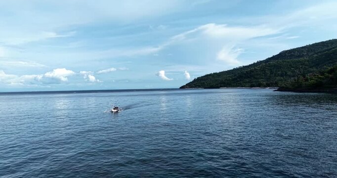 Drone aerial view of a traditional Balinese jukung fishing boat gliding across calm ocean waters near the lush tropical coastline of Amed, Bali, Indonesia beneath dramatic clouds.