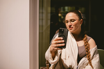 Fototapeta premium Smiling young woman with long braid and stylish glasses taking selfie on smartphone outdoors, happy female model posing for mobile photo or recording video blog in sunny European city street