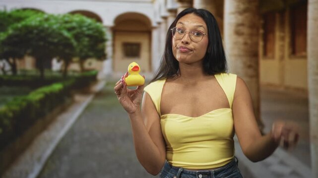Young hispanic brunette woman holds bright yellow rubber duck and points finger in stone building courtyard under arched colonnade; playful curiosity.