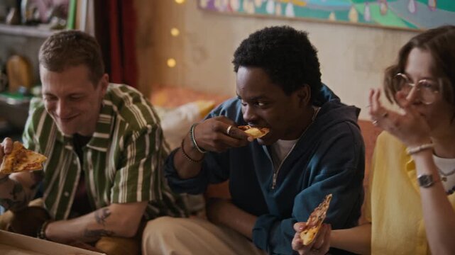 Medium long shot of three diverse young friends having fun and eating pizza while gathering at home