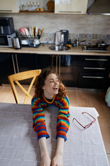 Fototapeta premium A young woman with curly hair wearing a colorful striped sweater smiles across a bright kitchen table, enjoying a casual home moment as sunlight streams in and sunglasses rest beside a laptop.