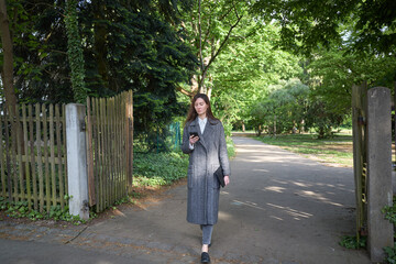 Fototapeta premium Woman in long coat walking in park and looking at her phone