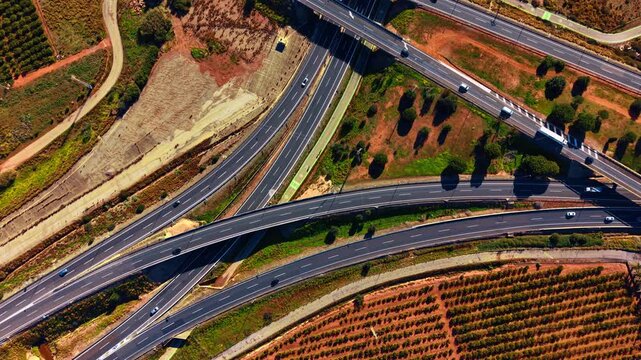 Top down view of complex highway interchange and orange groves. High angle shot of curved asphalt roads crossing red soil fields and green citrus plantations with moving vehicles.