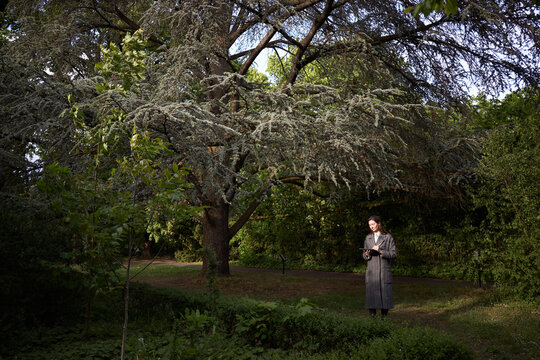Woman in long coat standing under a large tree in a park holding a tablet