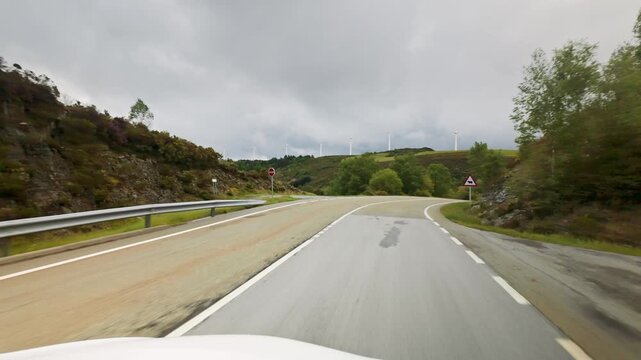 POV from a car moving along a winding road surrounded by green hills and trees in Asturias, Spain, on a cloudy day, with wind turbines visible in the distance generating renewable energy