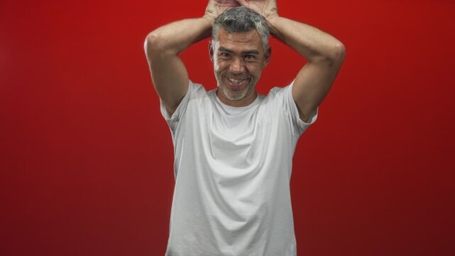 Man with grey hair in a white t shirt holding hands on head and smiling against a red studio backdrop; playful.