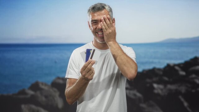 Man holding creditcard and smiling, covering right eye with hand gesture in studio; financial relief.