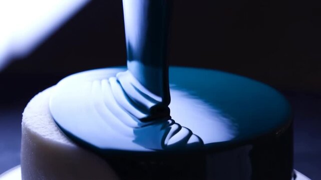 Slow motion close up of cake being glazed with dark blue glossy icing on studio table indoors