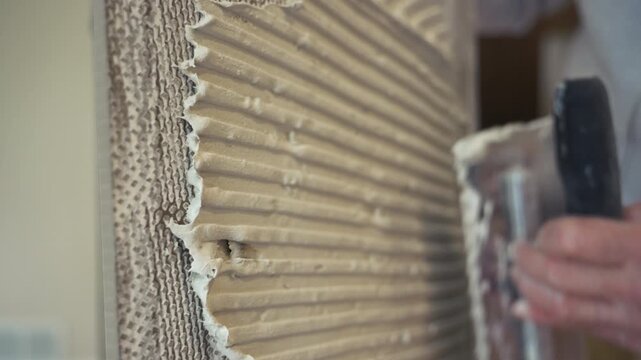 Professional worker applies thick layer of adhesive to ceramic tile to glue it to wall. renovation, building preparation, and construction tools 