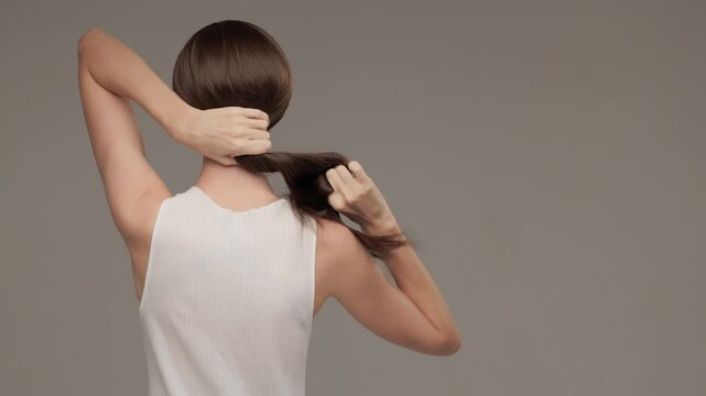The brunette gathers her long straight hair into a ponytail and twists it. View from behind. Copycpase