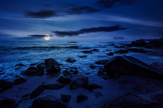 huge stones on a sandy beach at night. wonderful summer vacation season at the black sea. full moon over calm waves washing the sand shore. mysterious nature background with cloudy sky