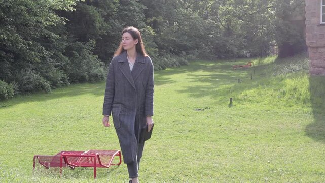 Woman in stylish coat walks through a sunny park with red benches