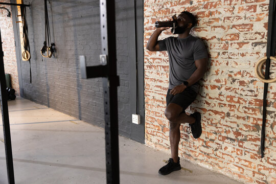 African American man in gray tee leaning on brick gym wall drinking from bottle, copy space