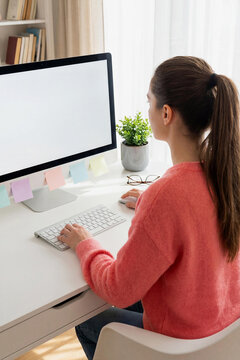 Rear view of a young Caucasian woman with a ponytail working on a computer at a white desk. Home office workspace with notes, a plant, and books. Concept for remote work, study, freelance lifestyle.