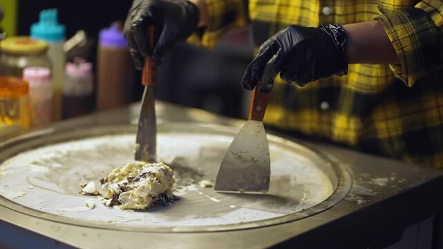 Rolled ice cream preparation with spatulas on cold plate at night market, Street food vendor mixing sweet cream and chocolate dessert 4k