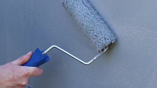 Painting a wall with a roller in gray. The process of applying gray paint to the surface using a paint roller. The master's hand applies a layer of gray paint to the wall using a textured paint roller