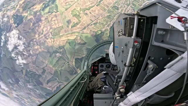 Aerobatic jet cockpit during steep roll above rural countryside, dramatic aviation video frame with farmland, clouds, open canopy and aircraft controls
