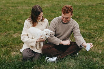 Obraz premium A couple sit on the grass outdoors, cuddling a white goat kid while a bottle feeds in a serene rural moment. A gentle, affectionate scene highlighting lifestyle, animal care, and family bonding in