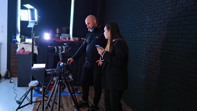 Videographer And Assistant Setting Camera In Studio. Man and assistant adjust camera on tripod near LED lights and black brick wall in studio.