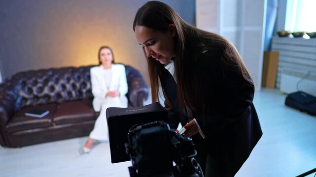 Assistant Adjusting Teleprompter Camera During Studio Interview. Assistant adjusts teleprompter on tripod camera while seated woman in white suit appears blurred behind.