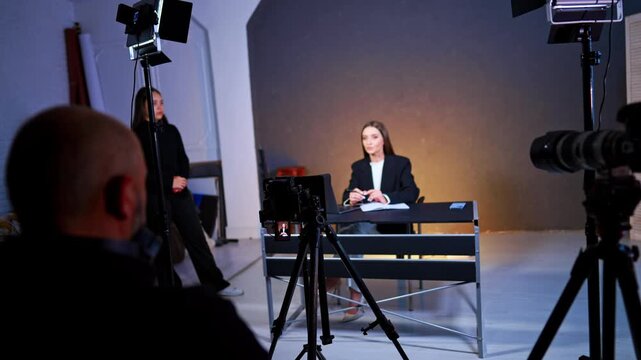 Professional video recording of woman at desk in studio. Wide studio view showing camera on tripod and lighting equipment while filming woman sitting at table.