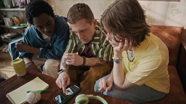 Zoom out shot of intercultural young friends checking food delivery menu on smartphone and discussing their order in rented apartment