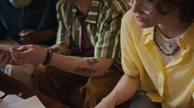Handheld shot of three diverse roommates sitting over table with past due bills and receipts and making calculations in rented apartment