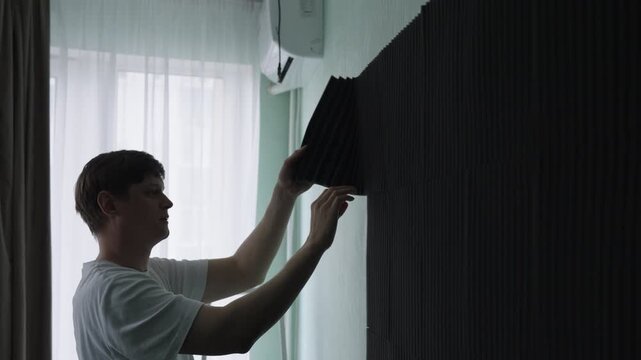 Focused handyman mounting dark polyurethane acoustic foam panels on wall, improving sound absorption, reducing echoes for better recording environment in professional studio. Shooting in slow motion.