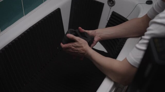 Man hands wringing out black acoustic foam panel in bathtub to remove excess water, showing material absorbent and flexible properties during cleaning and maintenance, close-up, slow motion.