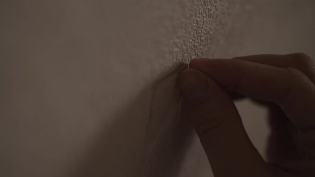 Close-up of fingers peel thin strip of flaking paint or wallpaper from textured wall. Detail shot conveys anxiety, boredom, deconstruction in dark interior of room. Shooting in slow motion.