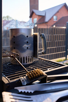 Smoking charcoal chimney starter stands on grill grate with barbecue brush and fork