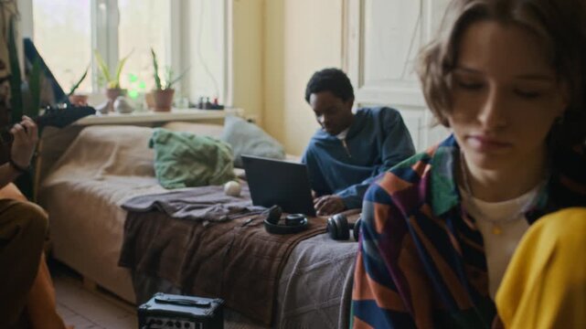 Handheld shot of young Caucasian woman using smartphone, male roommate practicing guitar chords, African American zoomer working on laptop in shared flat